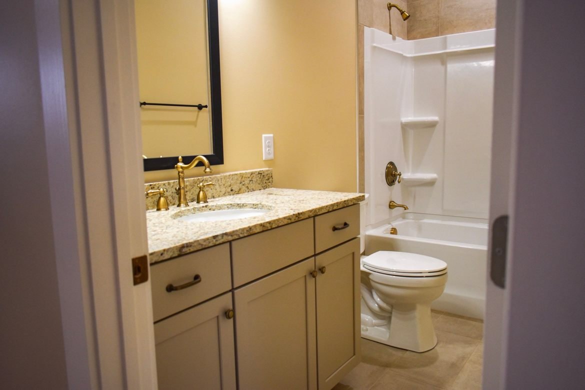 Bathroom with a single vanity, granite counter and brass faucet, and a tub with shower and toilet visible through the doorway